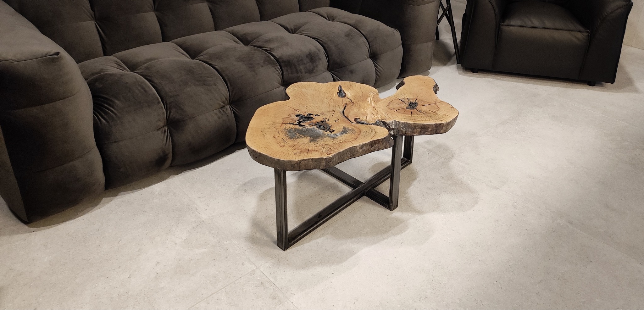 Niski stolik kawowy z blatem z naturalnego drewna o nieregularnym kształcie, wsparty na metalowej konstrukcji, ustawiony obok tapicerowanej sofy w odcieniach brązu i fotela.