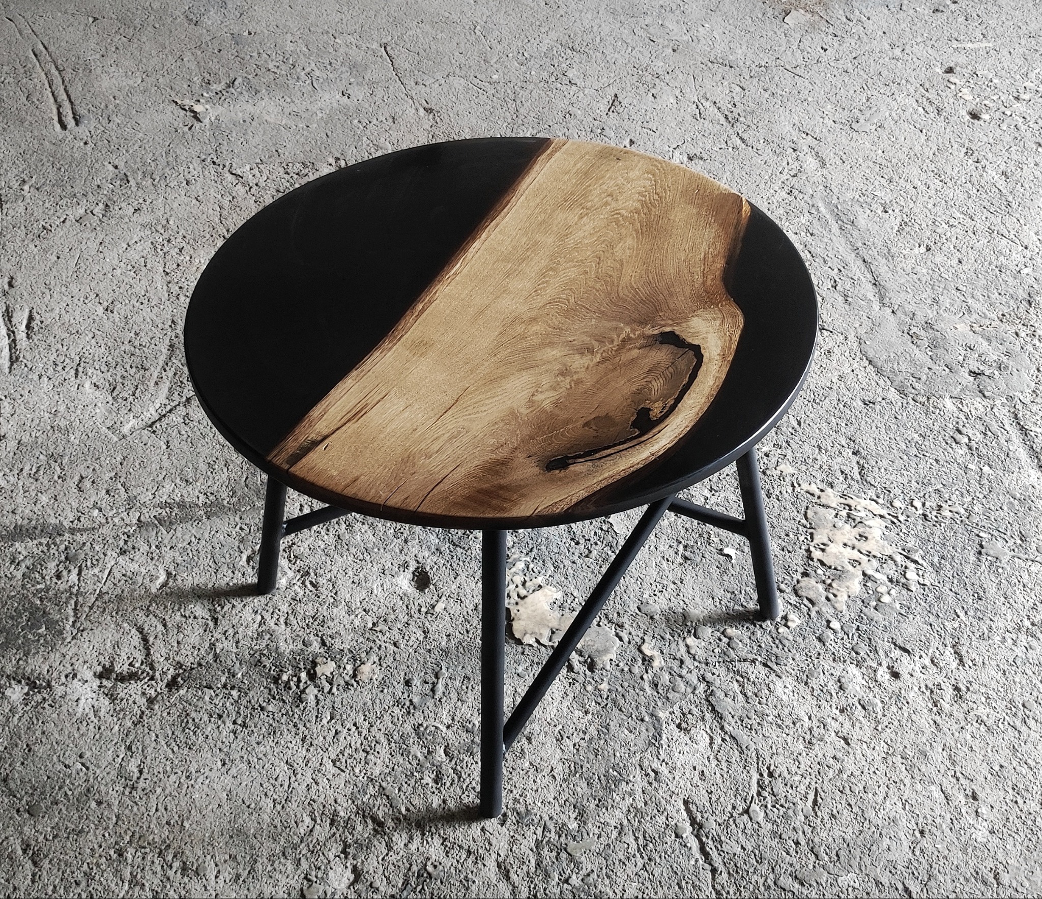 Okrągły stolik kawowy z blatem z drewna i czarnej żywicy epoksydowej, na metalowych, czarnych nogach, na surowym betonowym tle.
