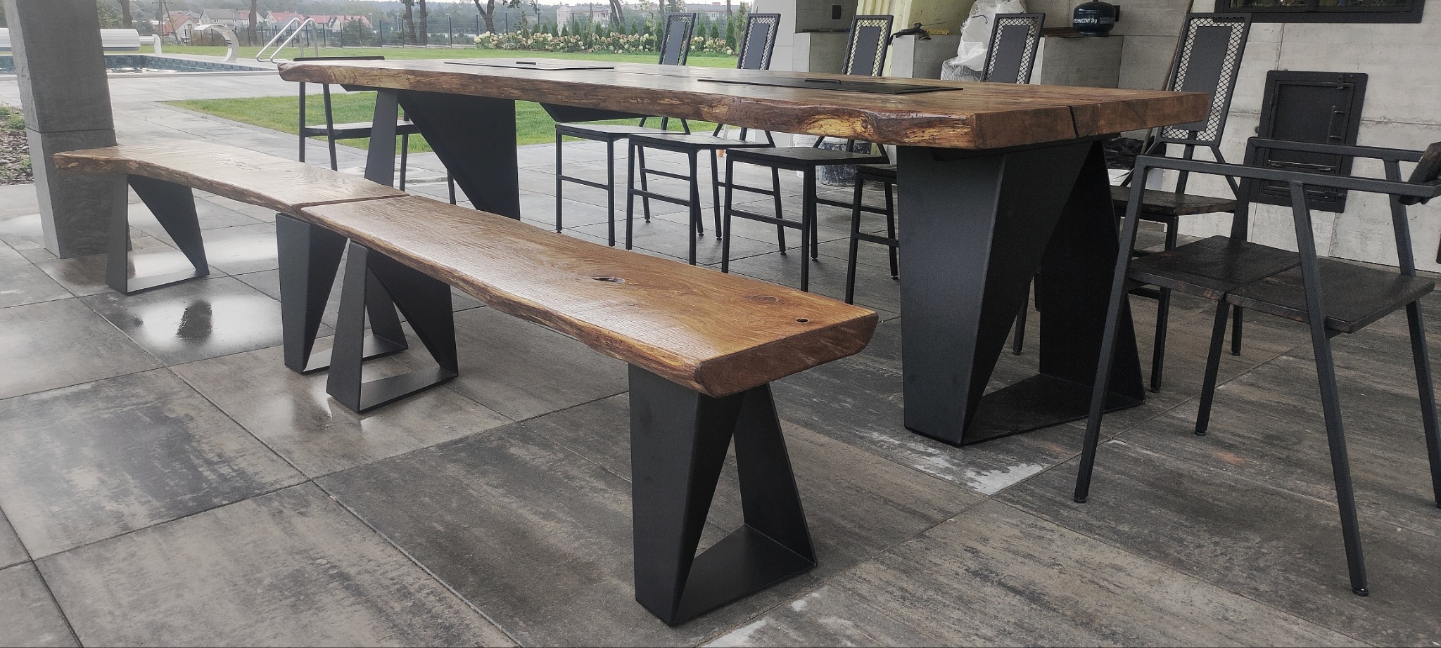 Zestaw mebli ogrodowych: stół z blatem z litego drewna o nieregularnych krawędziach, wsparty na geometrycznych, metalowych nogach, oraz ławka o podobnej konstrukcji, uzupełniony o metalowe krzesła,...