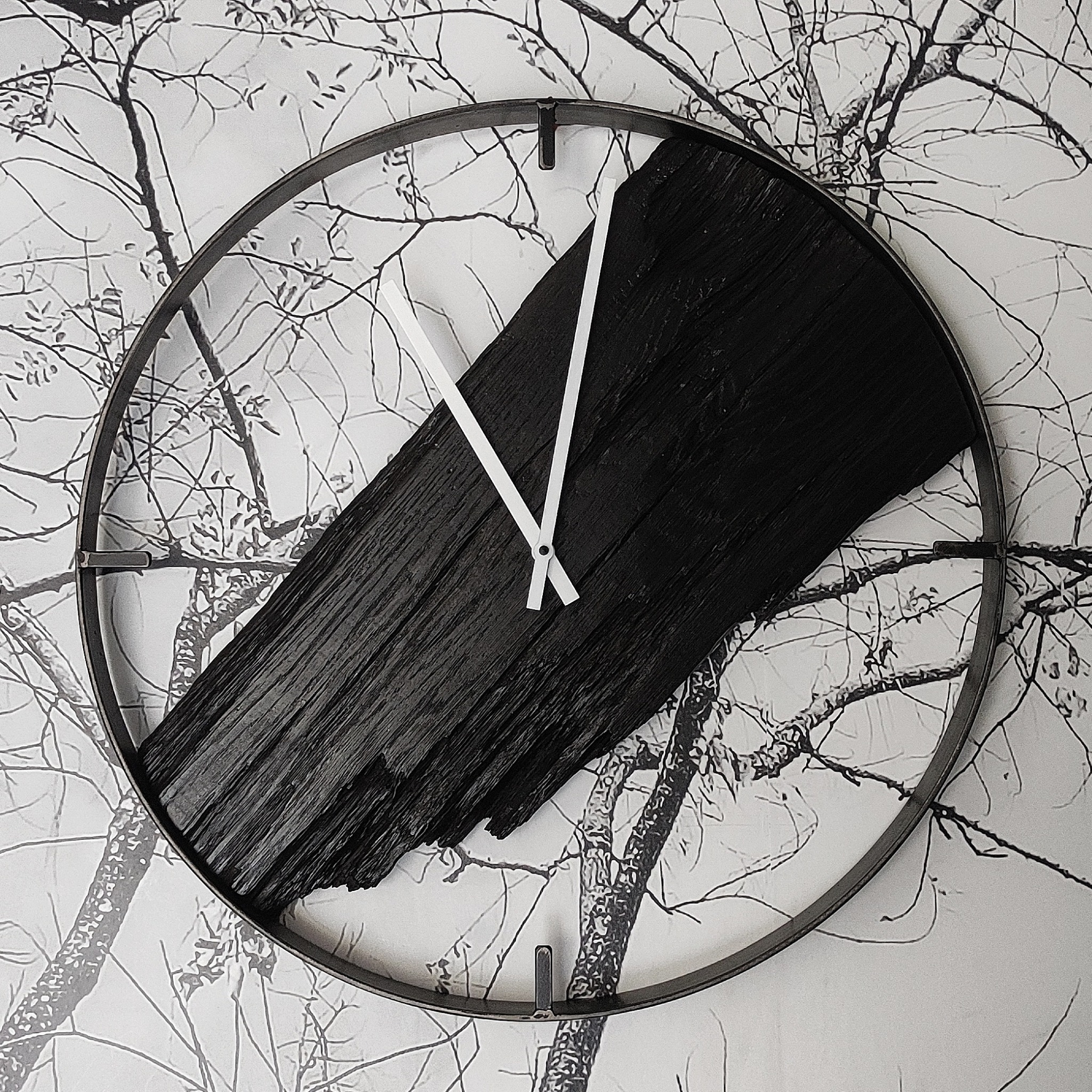 Okrągły zegar z metalową ramą, drewnianą tarczą w kolorze czarnym i białymi wskazówkami, zawieszony na ścianie z tapetą w motywie drzew bez liści.