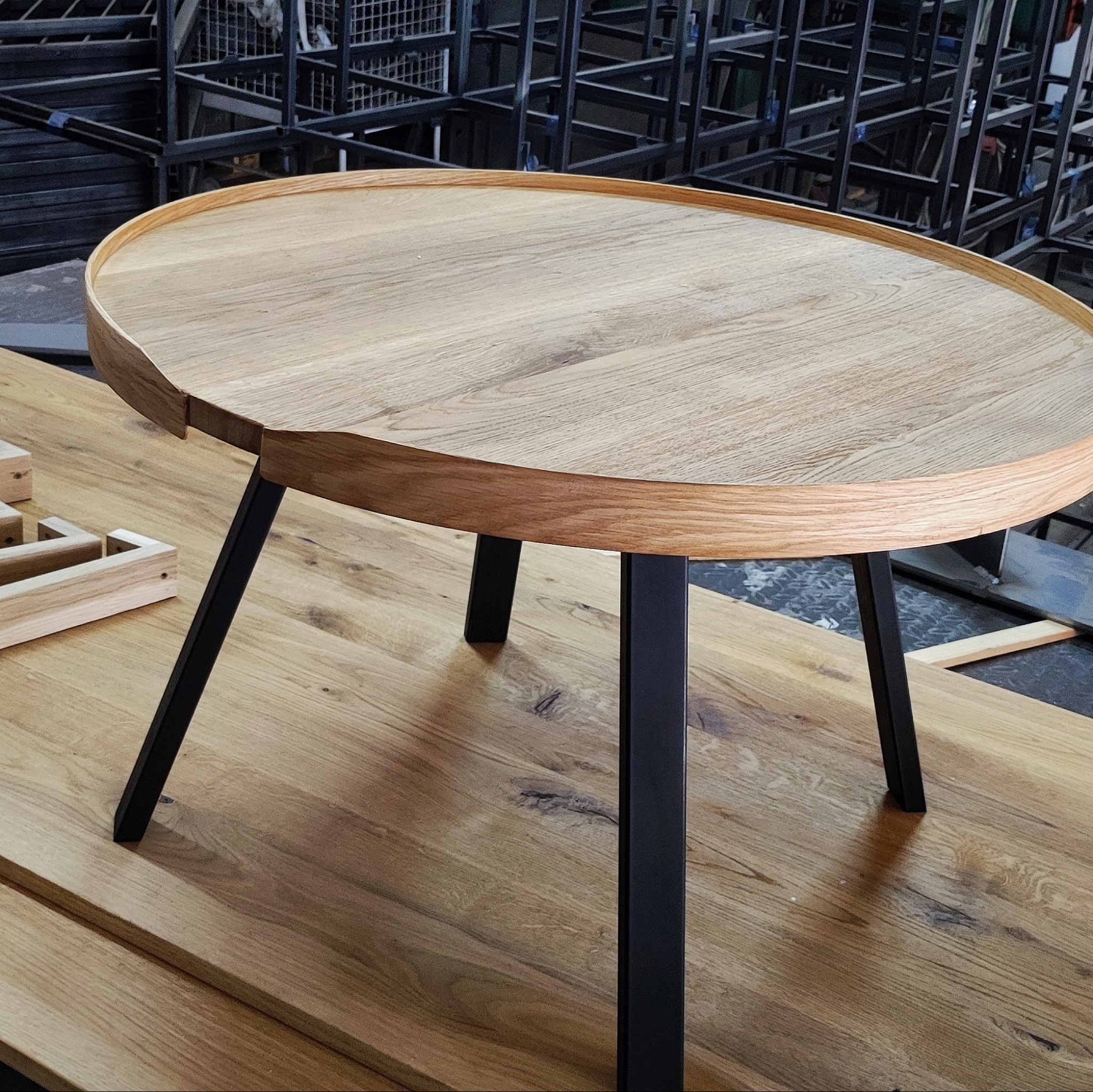 Okrągły stolik kawowy z blatem z jasnego drewna i czarnymi, metalowymi nogami, umieszczony na drewnianym podłożu; w tle widoczna konstrukcja z profili.