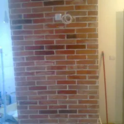 Ściana z czerwonej cegły z widocznymi fugami, gniazdko elektryczne z wystającymi kablami, fragment podłogi i ściany w tle.