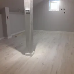 Wykończone wnętrze poddasza z jasnymi panelami podłogowymi i szarą ścianą, widoczny słup konstrukcyjny z puszkami elektrycznymi i małe okno dachowe.