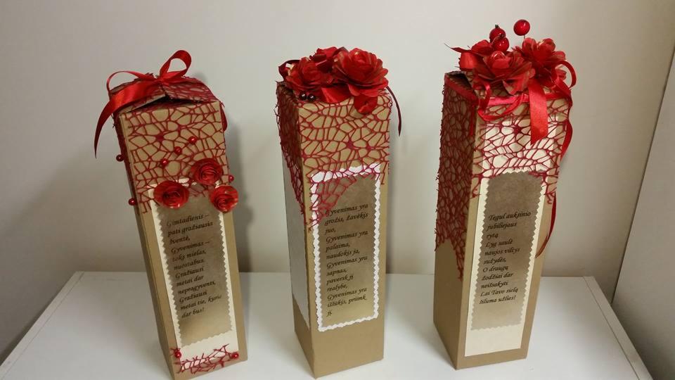 Trzy wysokie, ręcznie dekorowane pudełka prezentowe z czerwonymi wstążkami, siatką i papierowymi różami, z etykietami z tekstem w obcym języku, ustawione na białym blacie.