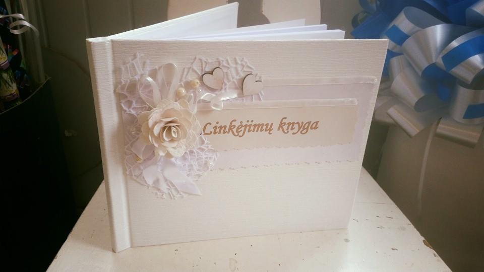 Ozdobny, biały album na życzenia z napisem 'Linkėjimu knyga', dekorowany koronką, różyczką i drewnianymi serduszkami, widoczny pod kątem na białym stole.