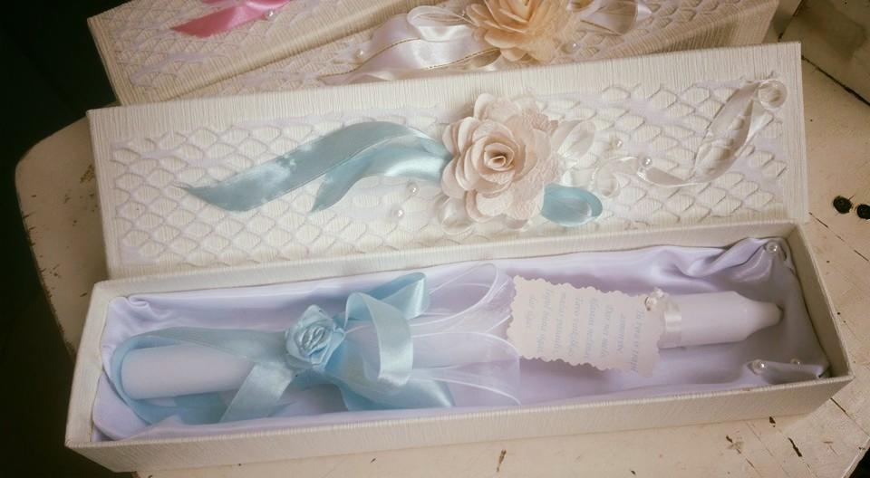 Dekoracyjna, biała skrzynka z teksturowanym wzorem, wypełniona białym materiałem i przedmiotami owiniętymi w białe i błękitne wstążki, zwieńczonymi papierową różą; na wieczku skrzynki ozdobny pas...