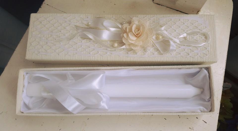 Otwarty, kremowy, teksturowany kartonik z dekoracją kwiatową i perłami na wieczku, wewnątrz biała świeca z satynową wstążką na białym atłasie.