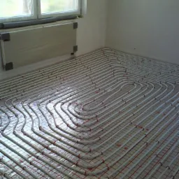 Białe rurki ogrzewania podłogowego ułożone na czerwonej siatce, widoczne na tle okna i grzejnika ściennego w jasnym pomieszczeniu.