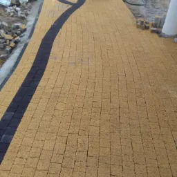 Żółta kostka brukowa ułożona w łukowaty wzór z czarnymi akcentami, częściowo ukończona, z widocznymi zapasami kostki i osobą w tle.