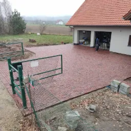 Ułożona czerwona kostka brukowa przed domem z garażem, widoczna brama wjazdowa z siatką i elementy do wykończenia, w tle drzewa i zabudowania.