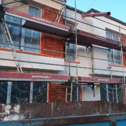 Dwupiętrowy dom w trakcie wykonywania elewacji z rusztowaniem, okna zabezpieczone folią, fragment ściany z cegły klinkierowej