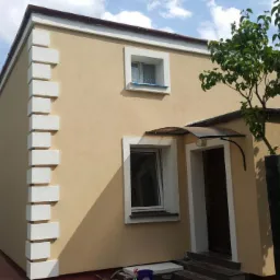 Ocieplony dom jednorodzinny z beżową elewacją, białymi detalami architektonicznymi, widok z boku na narożnik budynku, podbitka w kolorze brązowym.