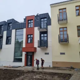 Nowo wybudowany budynek wielorodzinny z balkonami, widoczni dwaj mężczyźni zamiatający chodnik przed wejściem, teren wokół budynku częściowo zagospodarowany, częściowo w stanie surowym.