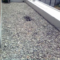 Płaski dach pokryty drobnymi szarymi kamieniami, z widocznym kablem elektrycznym zwiniętym w kłębek i biegnącym wzdłuż powierzchni, obok betonowej attyki i ściany budynku.