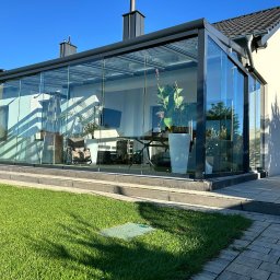 AlKac - Nowoczesna, przeszklona weranda z czarną ramą, przylegająca do jasnego domu. Widoczny trawnik i fragment chodnika z kostki. Wnętrze werandy wypełnione roślinami i meblami.