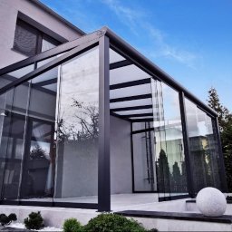 AlKac - Nowoczesna, przeszklona weranda z czarną ramą, przylegająca do jasnego budynku. Widoczne elementy ogrodu i błękitne niebo. Minimalistyczny design.