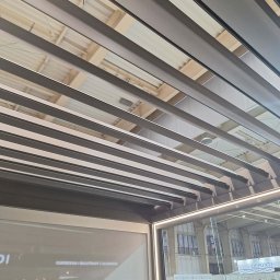 AlKac - Nowoczesna pergola z aluminiowymi lamelami, widok z dołu, z elementami ekspozycji targowej w tle. Minimalistyczny design, równoległe linie, sufit z widoczną konstrukcją.