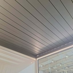 AlKac - Nowoczesna pergola tarasowa z aluminiowymi lamelami, widok od dołu, z oświetleniem LED i szklaną ścianą boczną. Widoczne odbicie otoczenia w szkle.