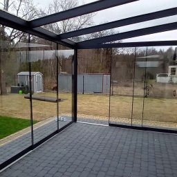 AlKac - Zadaszony taras z szarą kostką brukową, przeszklonymi ścianami i czarną konstrukcją stalową. Widok na ogród z domkami narzędziowymi w tle.