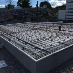 Zbrojona płyta fundamentowa z szalunkiem, widok z góry, przygotowana do zalania betonem, na tle ziemi i drzew pod błękitnym niebem.