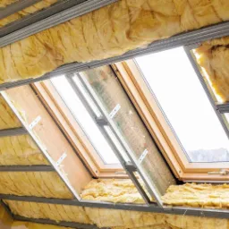 Izolacja poddasza wełną mineralną, widoczne metalowe profile konstrukcyjne i okno dachowe.