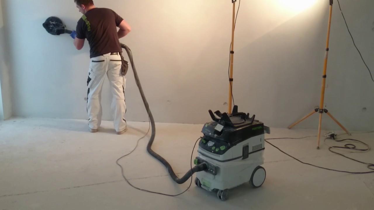 Szlifierka do gładzi podłączona do odkurzacza przemysłowego Festool, pracownik w trakcie szlifowania ściany w pustym pomieszczeniu, widoczne oświetlenie robocze.