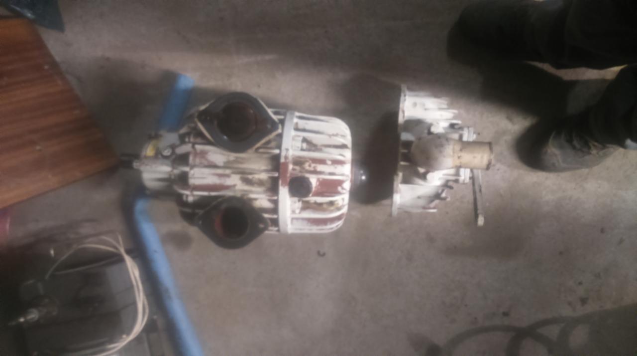 Rozłożona pompa hydrauliczna z widocznymi śladami użytkowania i resztkami farby, leżąca na betonowej podłodze obok narzędzi i fragmentu drewnianej płyty.