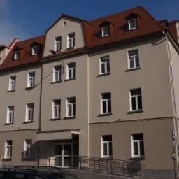 Odnowiony budynek biurowy z ceglastoczerwonym dachem i jasną elewacją, widoczny pod kątem z chodnika z barierkami dla niepełnosprawnych.