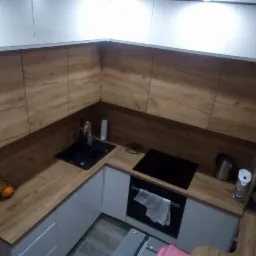 Nowoczesna kuchnia z drewnianymi blatami i frontami szafek w odcieniach brązu, połączona z białymi szafkami wiszącymi i dolnymi, czarny zlew i płyta indukcyjna, widok z góry na narożnik kuchni.