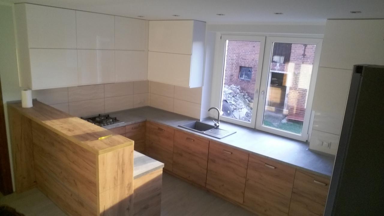 Nowoczesna kuchnia z białymi szafkami wiszącymi i drewnopodobnymi szafkami dolnymi, szary blat, okno z widokiem na ceglany budynek w tle.