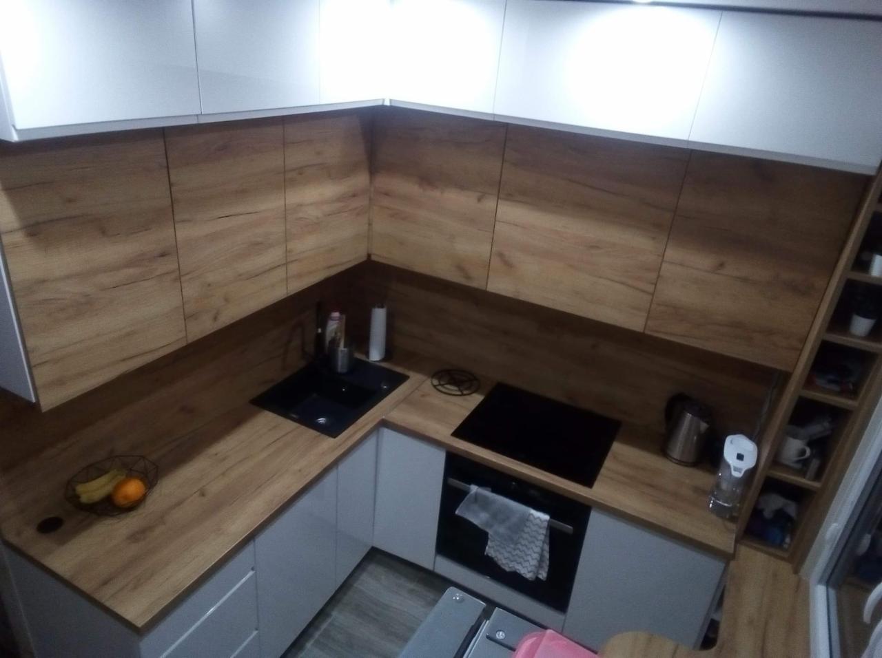 Nowoczesna kuchnia z drewnianymi blatami i frontami szafek w odcieniach brązu, połączona z białymi szafkami wiszącymi i dolnymi, czarny zlew i płyta indukcyjna, widok z góry na narożnik kuchni.