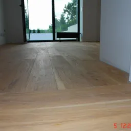 Jasna, drewniana podłoga ułożona w nowym domu, widoczne progi między pomieszczeniami i duże okno balkonowe z widokiem na zieleń.