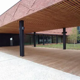 Zadaszone przejście z drewnianą podłogą i sufitem lamelowym w Łodzi, wsparte na czarnych, metalowych słupach, z ceglaną dekoracją elewacji.