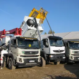 Trzy ciężarówki budowlane zaparkowane na placu: betonomieszarka Volvo z czerwoną pompą, wywrotka z podniesioną żółtą skrzynią ładunkową i biała ciężarówka Renault, wszystkie z logo firmy.