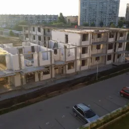 Wielopiętrowy budynek w trakcie budowy z białych bloczków, widok z góry na parking z samochodami, w tle bloki mieszkalne.