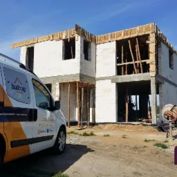 Budowa dwukondygnacyjnego domu z białych bloczków, widoczny samochód firmowy z logo Budovip oraz betoniarka, konstrukcja dachu w trakcie montażu, podpora z drewnianych bali.