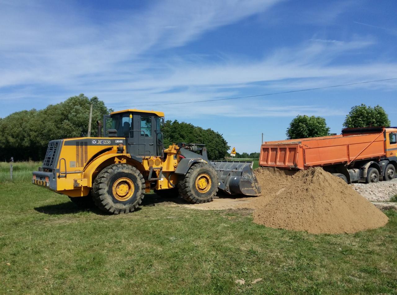 Żółta ładowarka kołowa XCMG LW640G obok pomarańczowej wywrotki z piaskiem na trawiastym terenie w słoneczny dzień.