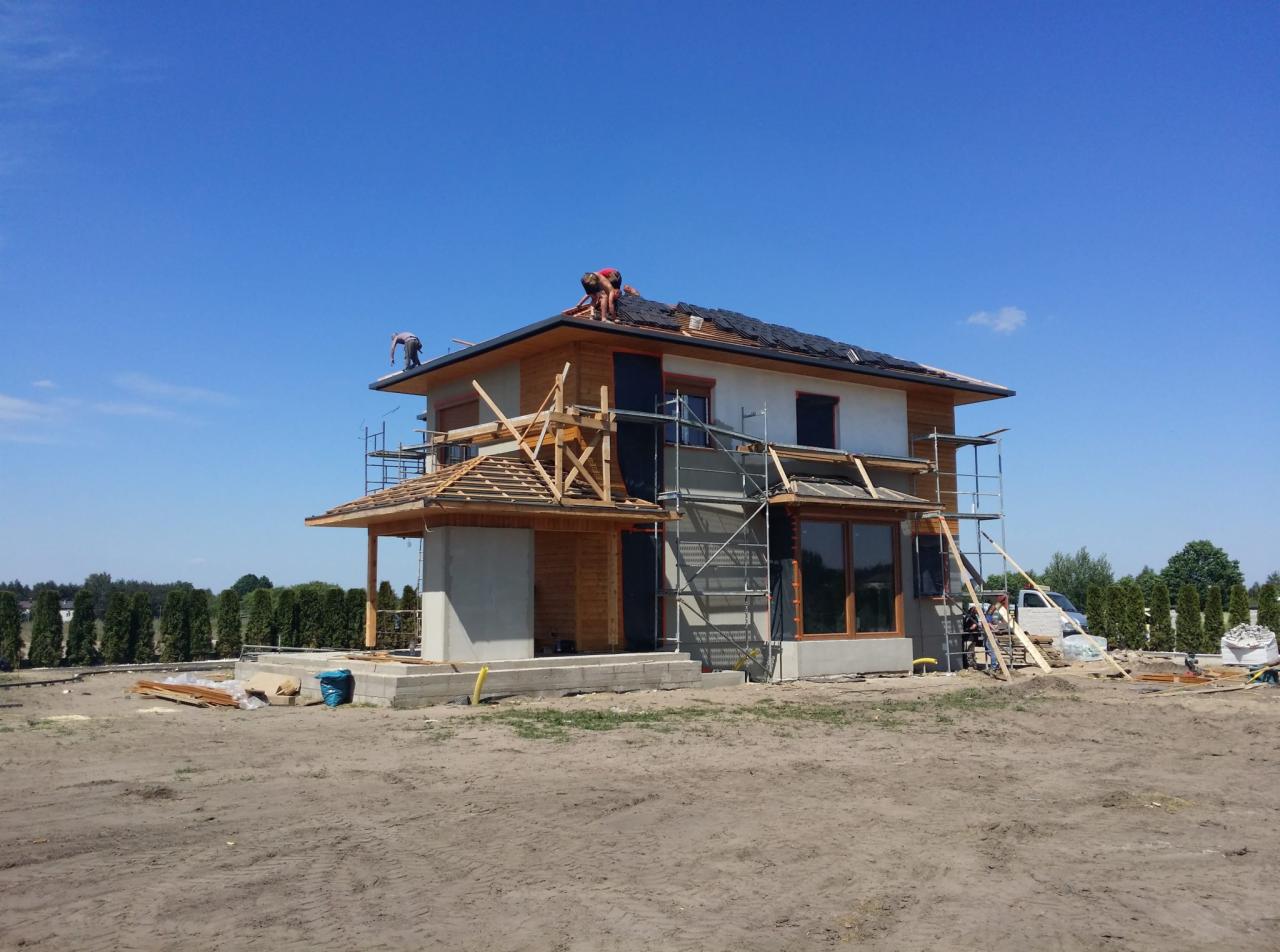 Dwukondygnacyjny dom w trakcie budowy, z widocznymi rusztowaniami, częściowo wykończoną elewacją drewnianą i pracownikami układającymi pokrycie dachowe na górze.