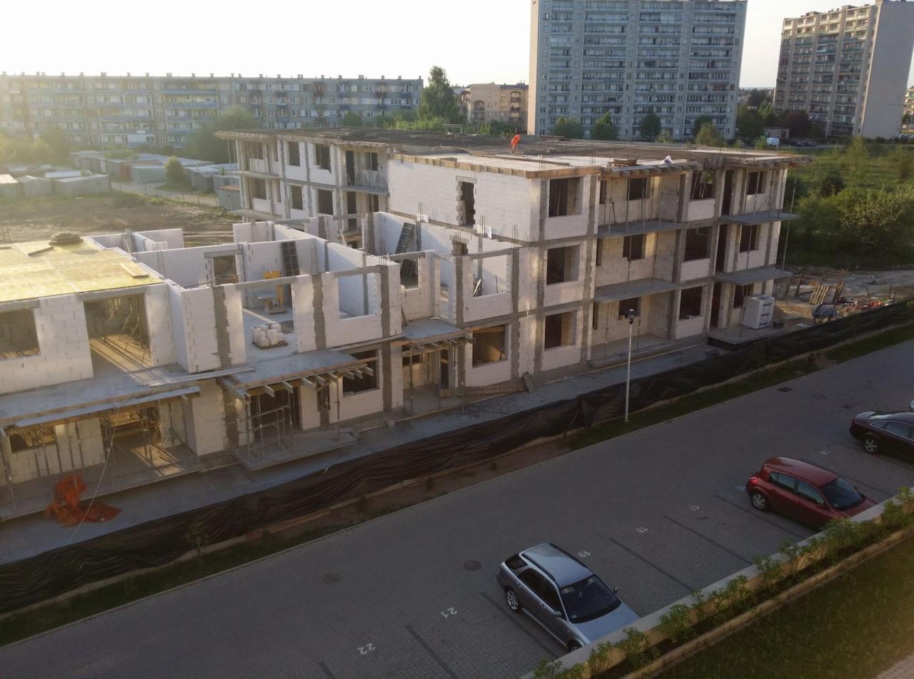 Wielopiętrowy budynek w trakcie budowy z białych bloczków, widok z góry na parking z samochodami, w tle bloki mieszkalne.