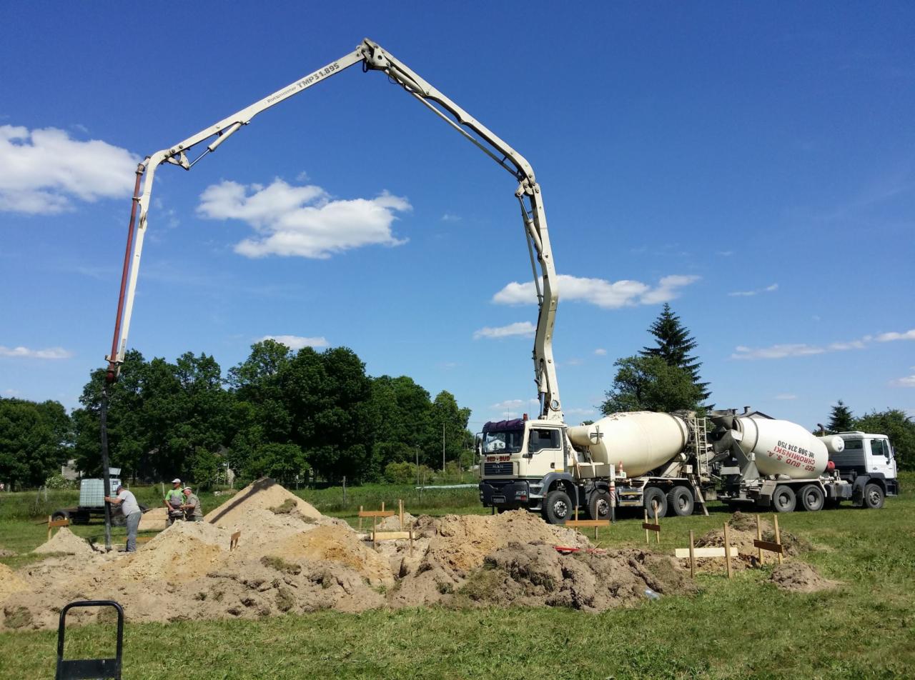 Wylewanie betonu z pompy do przygotowanego wykopu pod fundamenty budynku, widoczne dwa betonomieszarki i pracownicy budowlani.