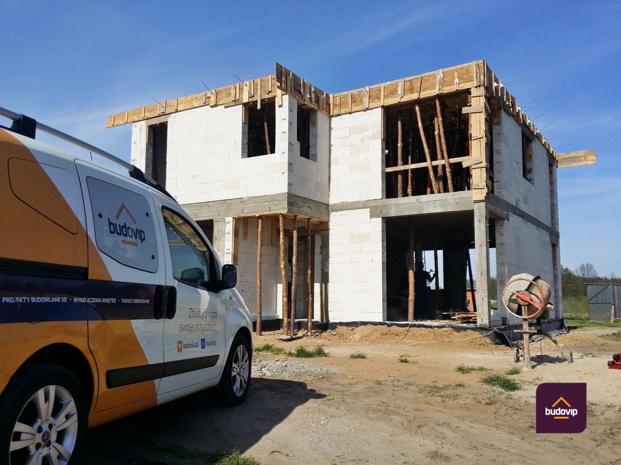 Budowa dwukondygnacyjnego domu z białych bloczków, widoczny samochód firmowy z logo Budovip oraz betoniarka, konstrukcja dachu w trakcie montażu, podpora z drewnianych bali.