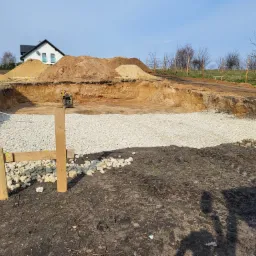 Wykop pod fundamenty domu jednorodzinnego z wysypaną warstwą gruzu, widoczne deski szalunkowe i ubijarka wibracyjna, w tle hałdy piasku i ziemi oraz dom w budowie.