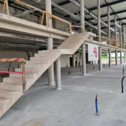 Surowe betonowe schody w surowym stanie wewnątrz budynku w trakcie budowy, z widocznymi kolumnami i instalacjami wystającymi z podłogi.