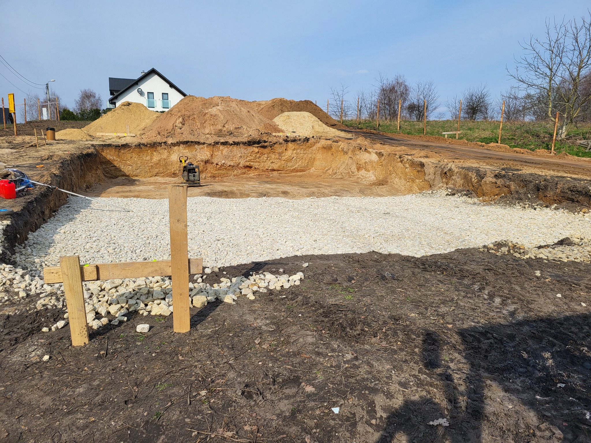 Wykop pod fundamenty domu jednorodzinnego z wysypaną warstwą gruzu, widoczne deski szalunkowe i ubijarka wibracyjna, w tle hałdy piasku i ziemi oraz dom w budowie.