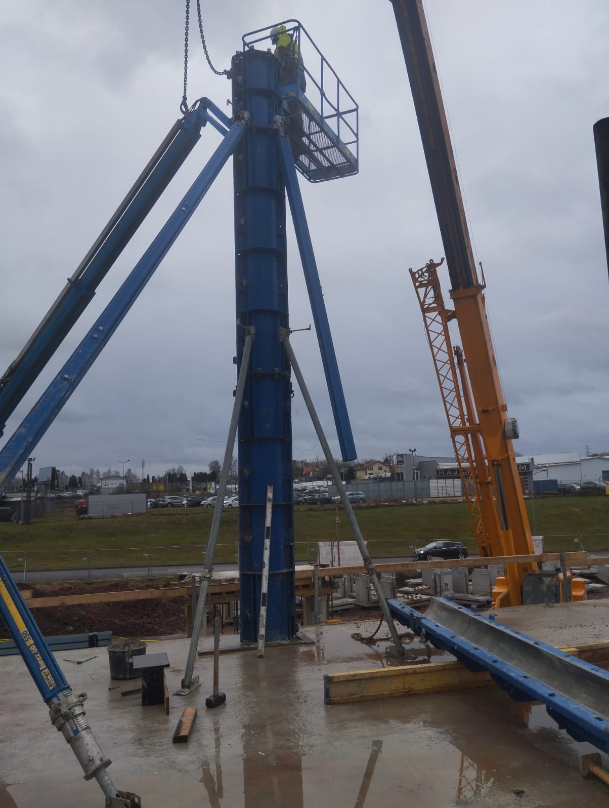 Montaż szalunku słupowego na mokrej płycie fundamentowej. W tle widoczny żuraw budowlany i zabudowa miejska.