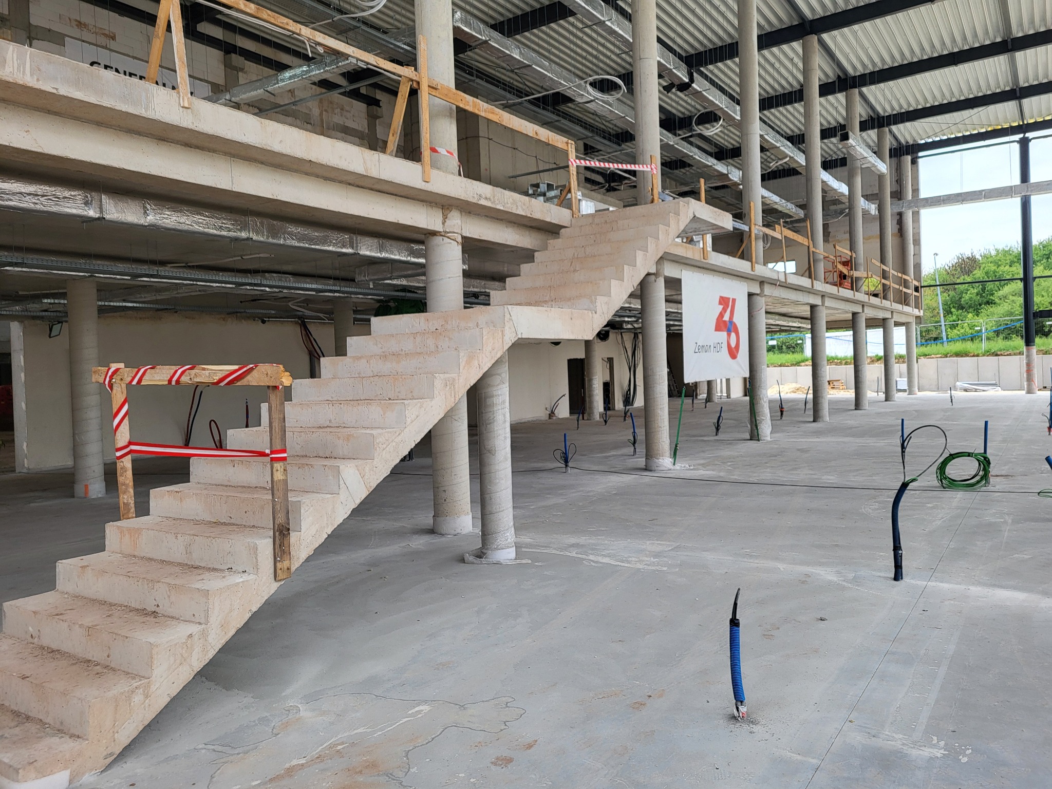Surowe betonowe schody w surowym stanie wewnątrz budynku w trakcie budowy, z widocznymi kolumnami i instalacjami wystającymi z podłogi.