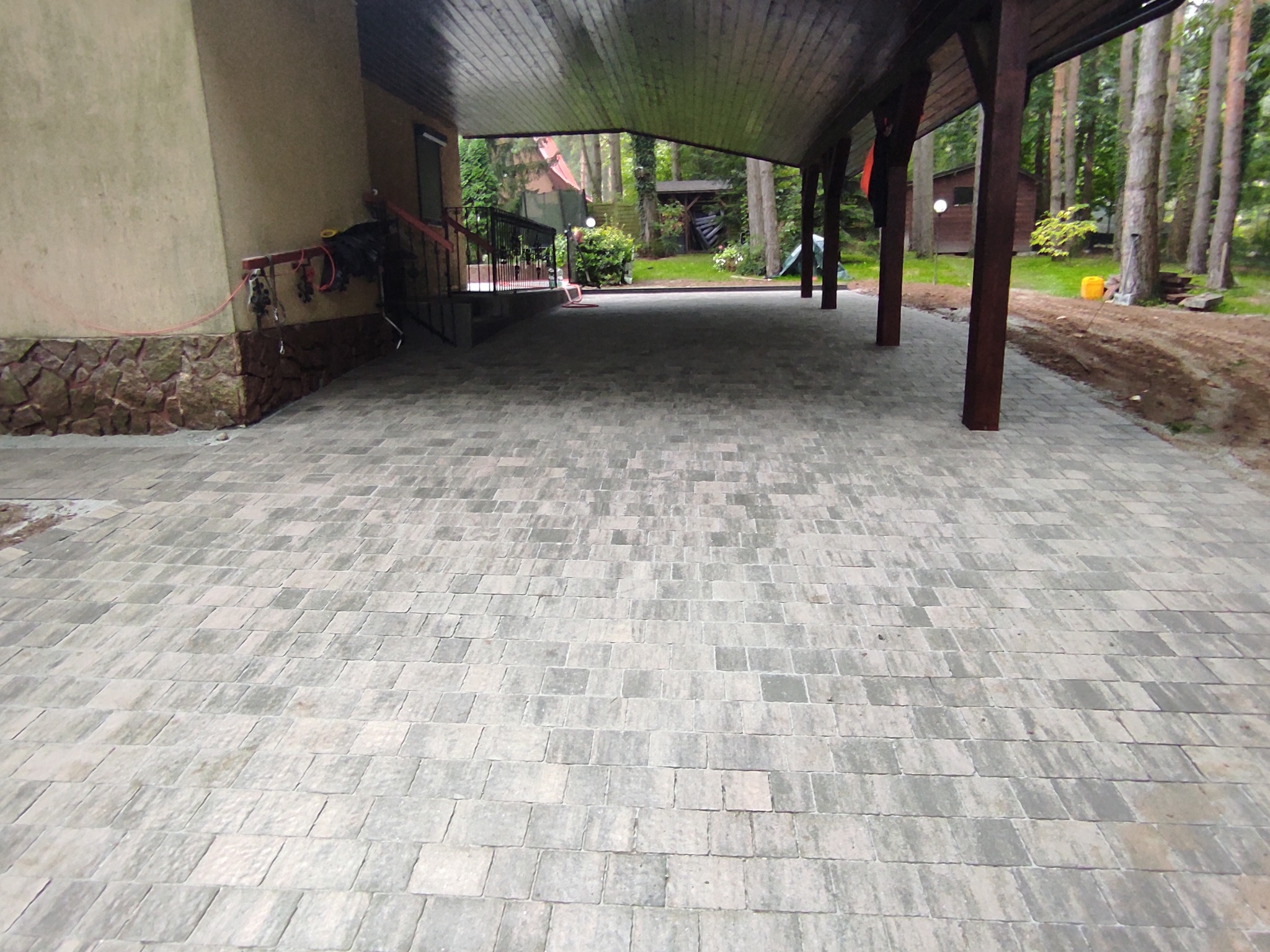 Nowa szara kostka brukowa ułożona pod zadaszeniem przylegającym do domu z kamienną elewacją, widok na ogród z drewnianymi domkami.