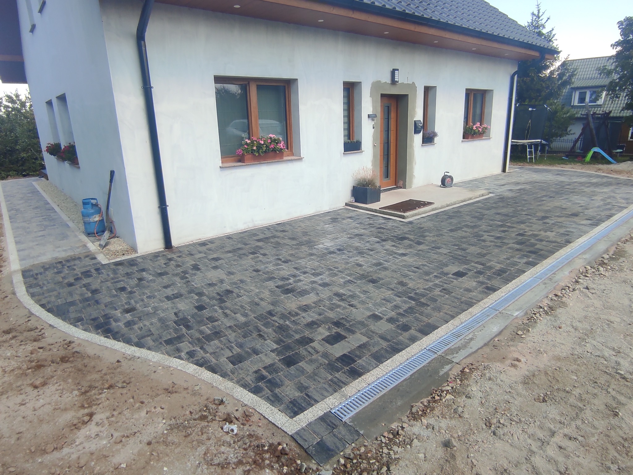 Ułożona kostka brukowa w kolorze grafitowym przed domem z białymi ścianami i drewnianą stolarką okienną, widoczny odpływ liniowy i gazowa butla w rogu budynku.