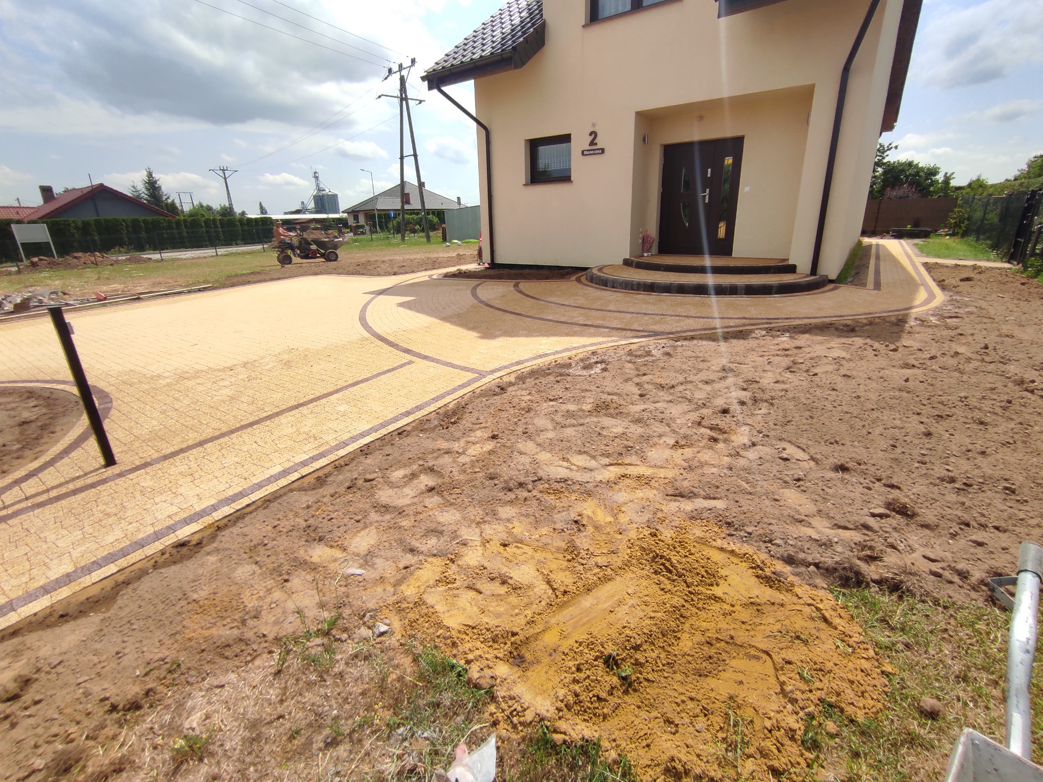 Żółta kostka brukowa ułożona w kształt podjazdu i chodnika wokół domu jednorodzinnego z ciemną stolarką okienną i drzwiową, widoczna sterta piasku i wózek transportowy w tle.
