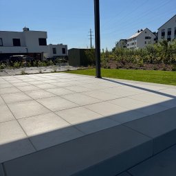 GreenPolis Sp. z o.o. - Nowoczesny taras z szarych płyt, widok na zadbany trawnik, domek narzędziowy i zabudowę jednorodzinną w tle, oświetlony słońcem.